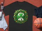 The Garment Graphics black graphic T-shirt features a bold green shamrock clover design in the center with smaller shamrocks and gold accents. Perfect for St. Patrick’s Day, its styled outdoors with a basketball, sports shorts, towel, and clear water bottle.