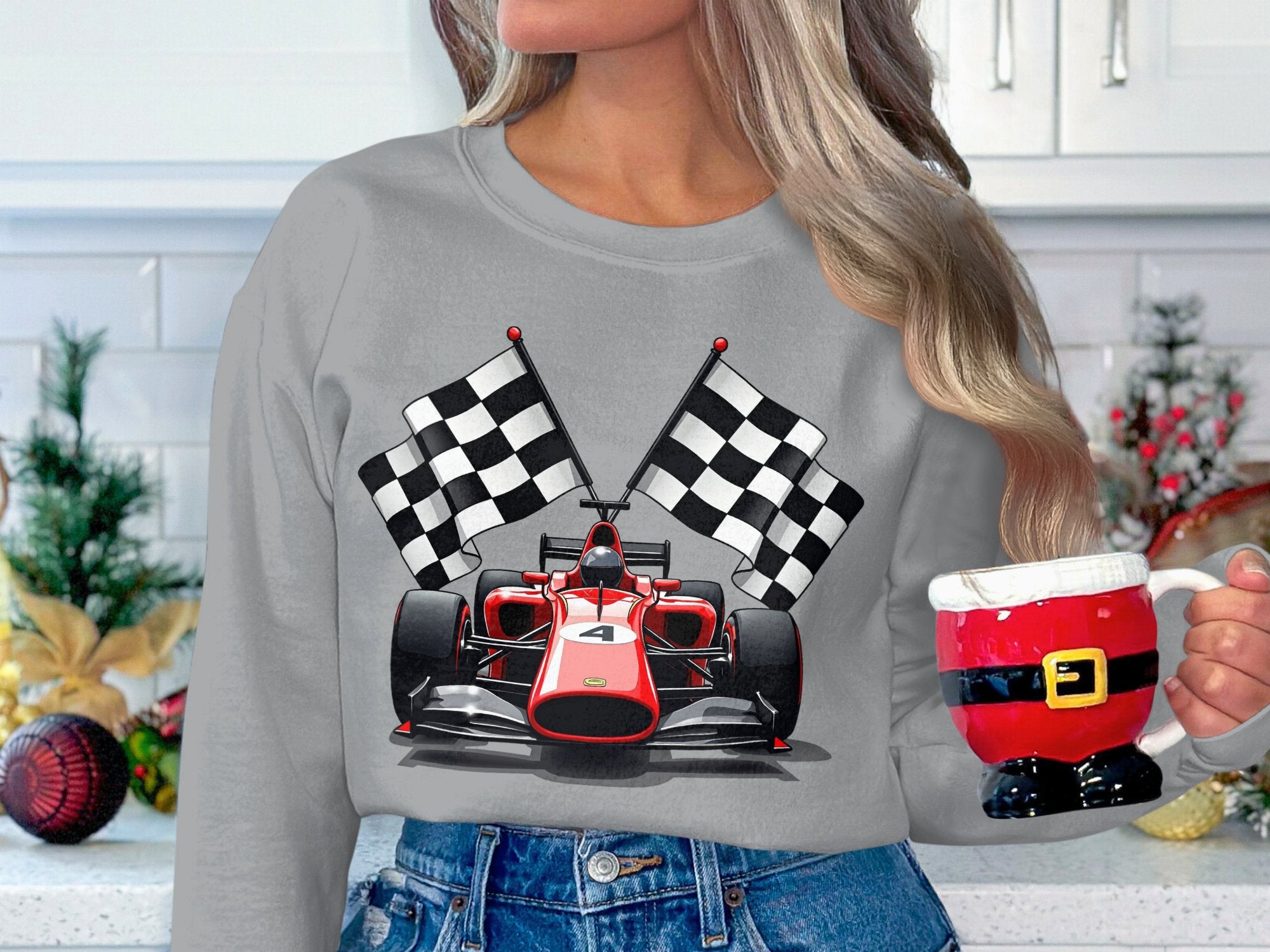 A person in a Garment Graphics gray sweatshirt with a racing car and checkered flags holds a Santa suit-themed mug. The backdrop is a holiday-decorated white kitchen, ideal for motorsport fans enjoying the festive season.