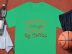 A green classic fit Garment Graphics T-shirt, featuring This day is powered by coffee in orange letters, is displayed on a basketball court beside a basketball, black shorts, a white towel, and a water bottle. Feel the energy with our Coffee T-Shirts collection.