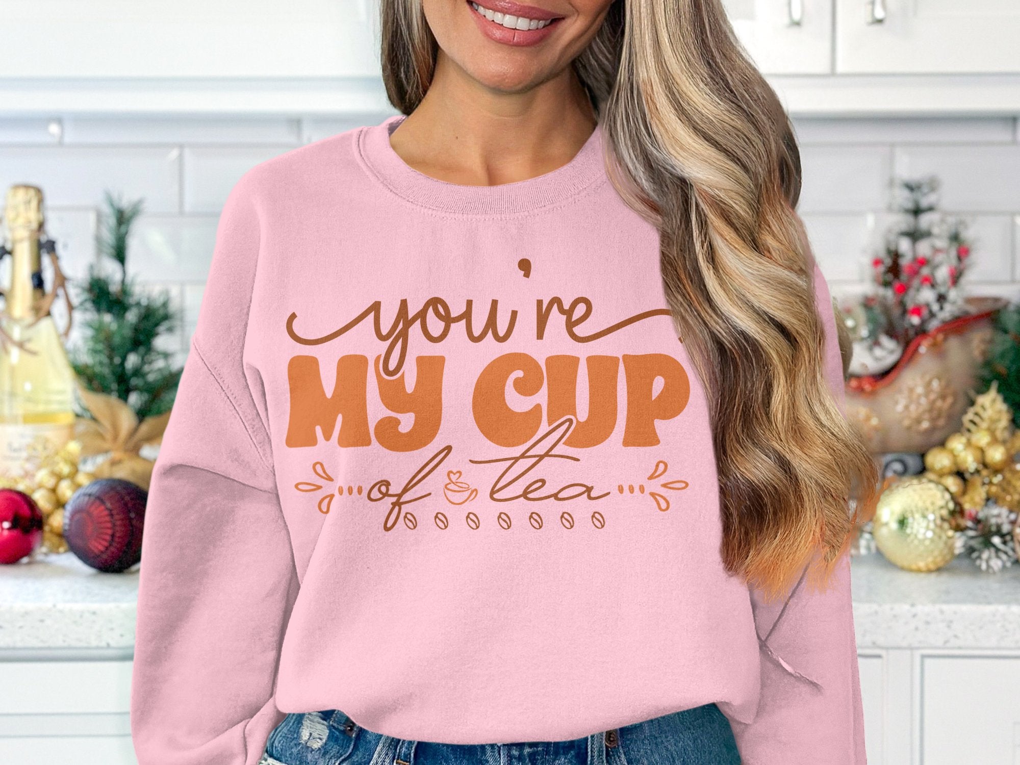 A person wearing a Garment Graphics unisex sweatshirt inscribed with youre my cup of tea in brown lettering stands in a festive kitchen, holding a cup.