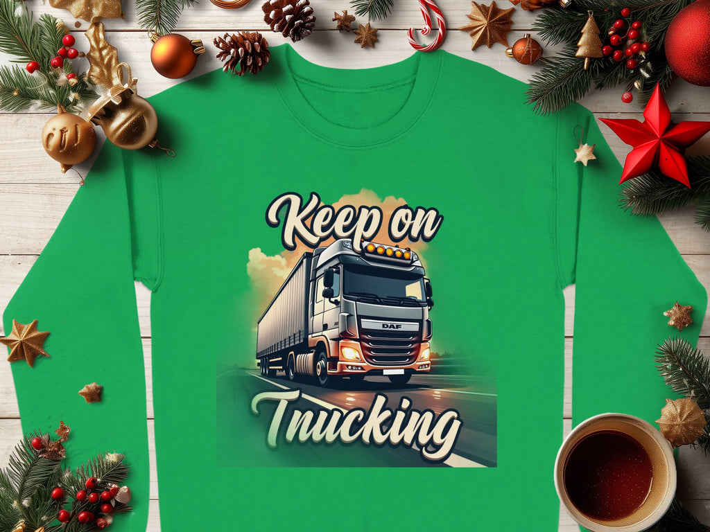 A green Graphic Inspirational Sweatshirt from Garment Graphics features a truck and Keep on Trucking text, set amidst festive decor like pine branches, ornaments, cinnamon sticks, and a steaming coffee cup on a wooden surface, capturing the open road spirit.