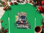 A green Graphic Inspirational Sweatshirt from Garment Graphics features a truck and Keep on Trucking text, set amidst festive decor like pine branches, ornaments, cinnamon sticks, and a steaming coffee cup on a wooden surface, capturing the open road spirit.