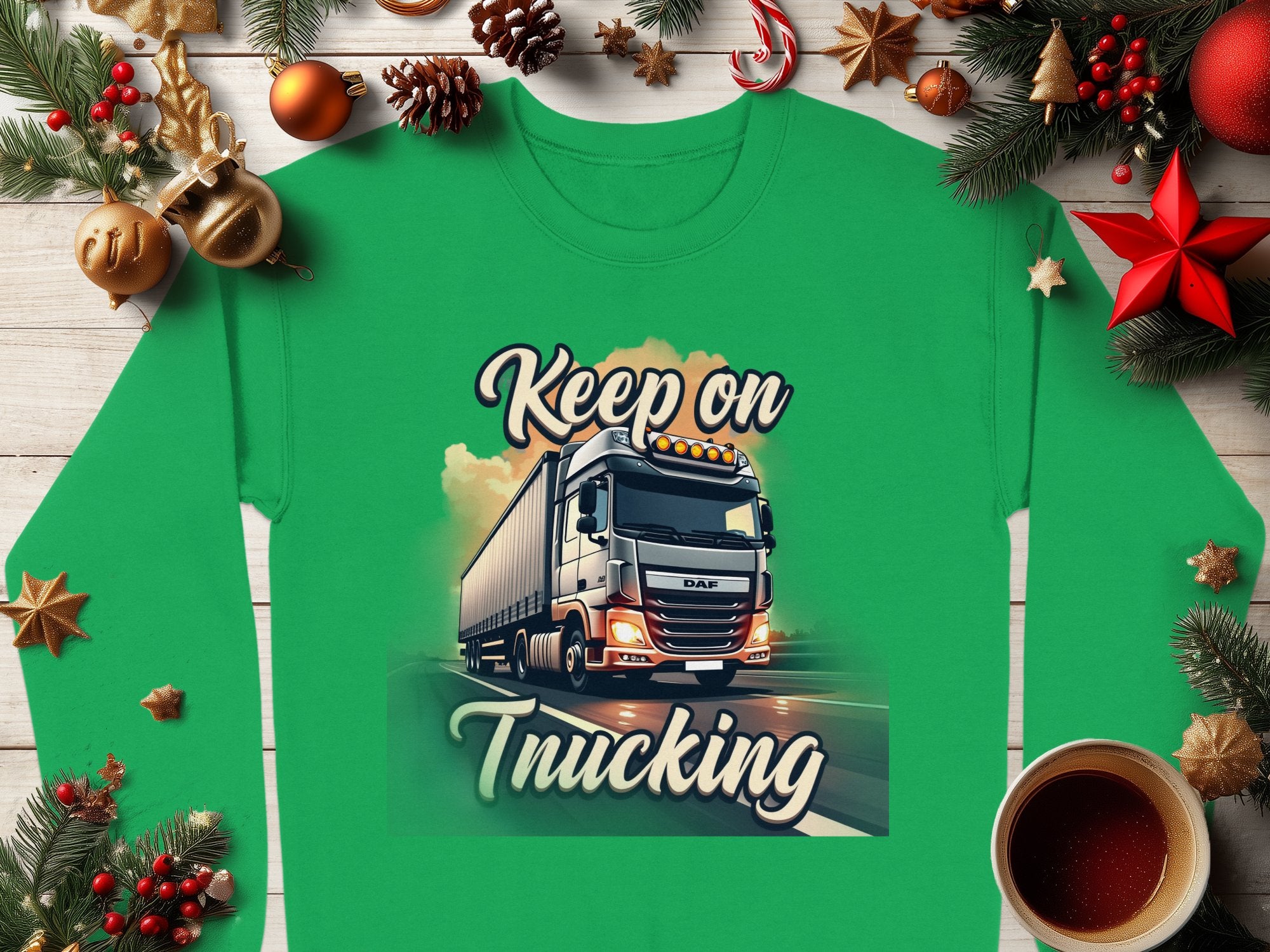 A green Graphic Inspirational Sweatshirt from Garment Graphics features a truck and Keep on Trucking text, set amidst festive decor like pine branches, ornaments, cinnamon sticks, and a steaming coffee cup on a wooden surface, capturing the open road spirit.