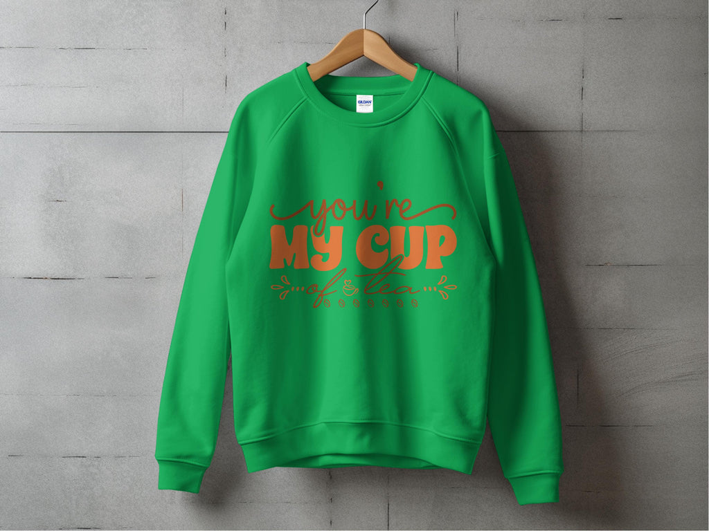 A cozy green sweatshirt by Garment Graphics, ideal for tea lovers, displays Youre My Cup of Tea in orange cursive on it. It hangs on a wooden hanger against a gray textured wall.