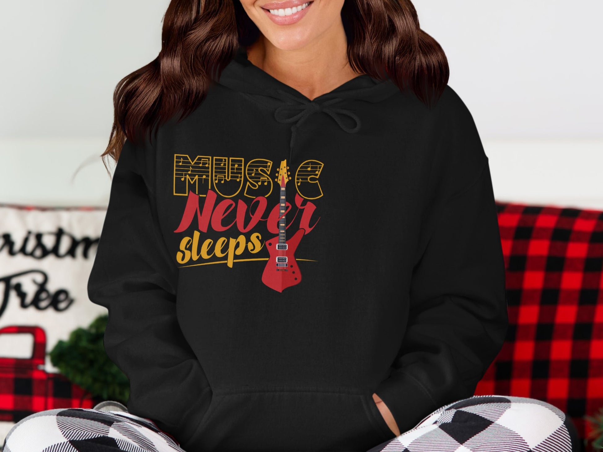 Someone is wearing a classic fit black hoodie from Garment Graphics with Music Never Sleeps and a guitar design. They are in a room with holiday decor, including a Christmas Tree sign and a cozy red and black plaid blanket.