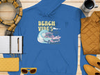 Garment Graphics vibrant blue hoodie, showcasing a Beach Vibes retro design with a vintage car, wave, and surfboards graphic, is laid on wood alongside a yellow jacket, shoes, sunglasses, cup, and an open notebook with pen.