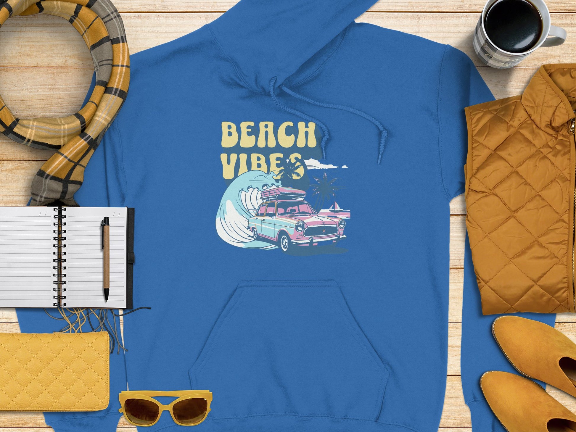 Garment Graphics vibrant blue hoodie, showcasing a Beach Vibes retro design with a vintage car, wave, and surfboards graphic, is laid on wood alongside a yellow jacket, shoes, sunglasses, cup, and an open notebook with pen.