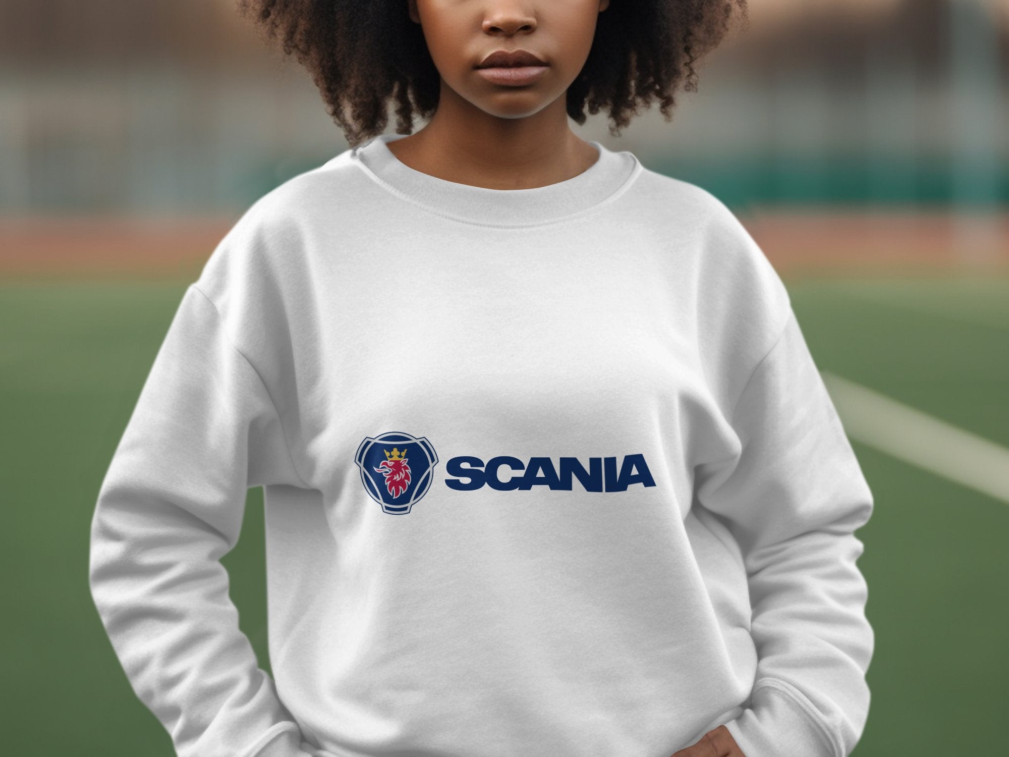 On a sports field, someone wears a unisex white sweatshirt from Garment Graphics featuring a prominent Scania logo, with the blurred background suggesting leisurely outings.