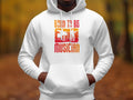 A man wears a Garment Graphics white unisex hoodie featuring Born to Be Musician above drummer and musician illustrations on a vibrant orange-red gradient, all set against a blurred autumn backdrop, embodying the spirit of music lovers.