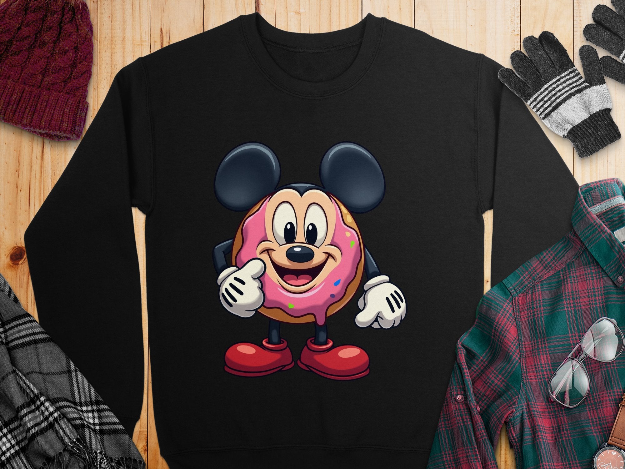 A black sweatshirt from Garment Graphics showcases a donut character with large round ears, white gloves, and red shoes. Paired with unisex beanie, gloves, scarf, and plaid shirt on a wooden surface.