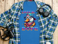 Garment Graphics Ducks Dope T-Shirt is a blue tee with a stylish duck in a hat, glasses, and scarf. Duck Dope is printed prominently. Its displayed on wood beside a plaid shirt, headphones, camera, and coffee cup, highlighting its unique style.