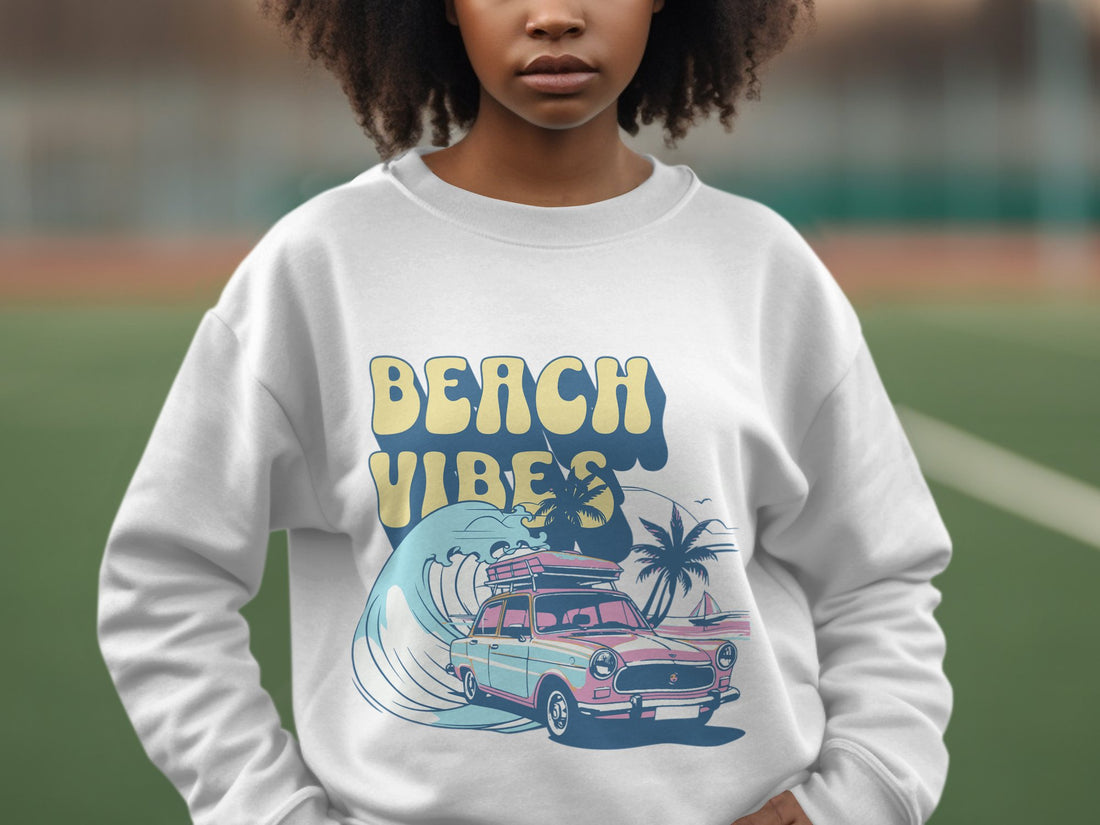 A person is wearing a Garment Graphics white sweatshirt with Beach Vibes in yellow and blue, featuring a pink classic car, surfboards, and palm trees. The retro design shines through the blurred background with greenery, ideal for beach fun.