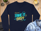 A navy blue sweatshirt from Garment Graphics unisex comfy casual collection is on a wooden surface. The yellow and turquoise Take It Easy Graphic with arrows and notes stands out. A folded denim shirt, a latte, and a small potted plant complete the scene.