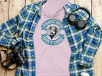 A pink Fishing Hair Dont Care hoodie by Garment Graphics lies on wood, accompanied by a blue plaid shirt, black headphones, camera, and a white lidded cup—ideal for anglers seeking style and comfort.
