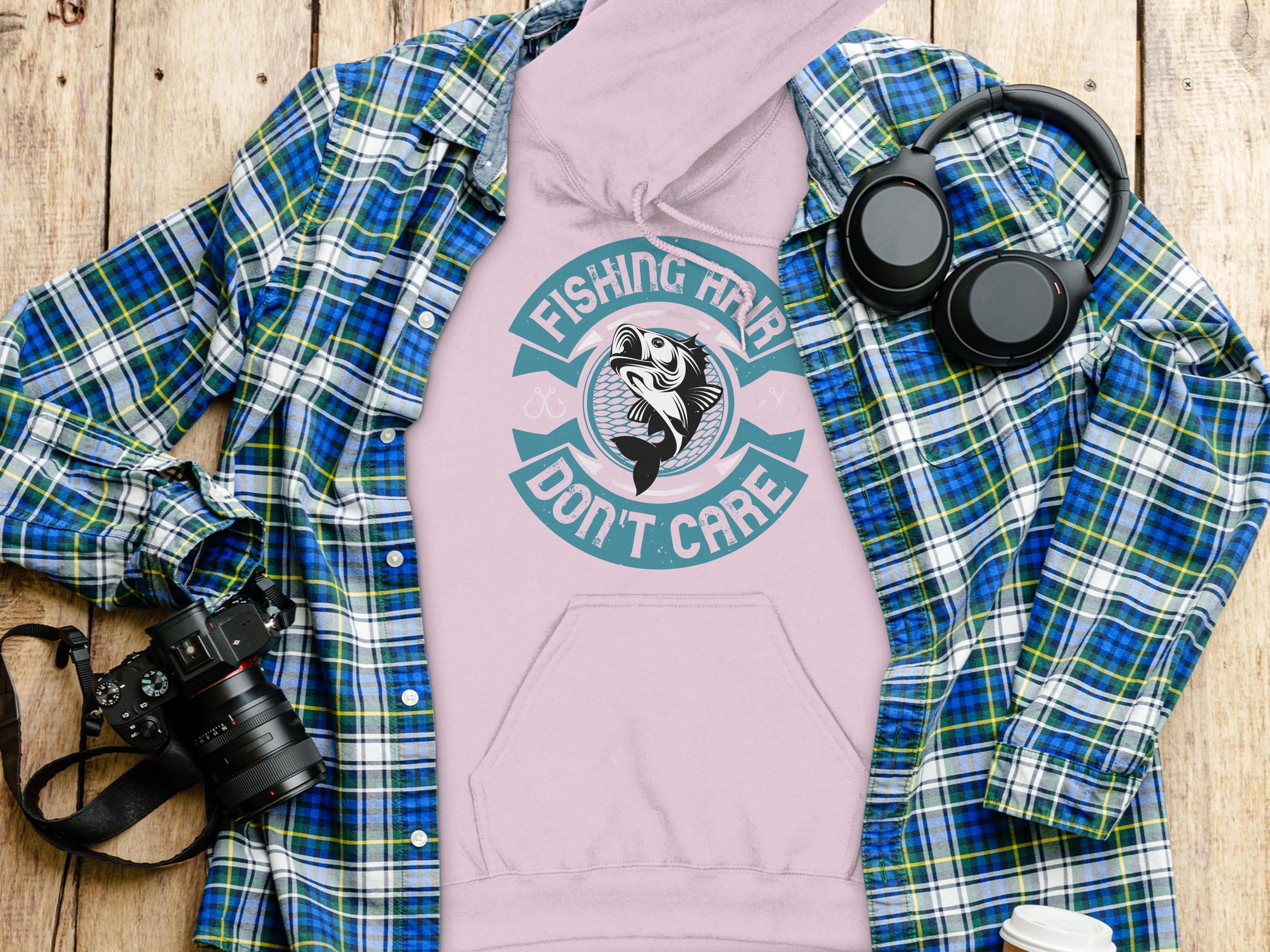 A pink Fishing Hair Dont Care hoodie by Garment Graphics lies on wood, accompanied by a blue plaid shirt, black headphones, camera, and a white lidded cup—ideal for anglers seeking style and comfort.