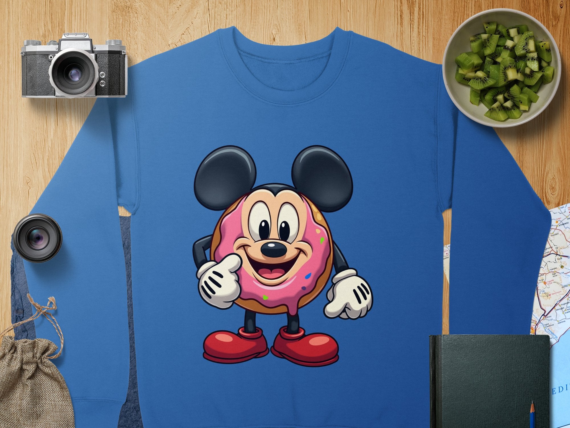 Garment Graphics blue unisex sweatshirt features a donut character reminiscent of a famous mouse in costume. Laid on wood and surrounded by a camera, lens, kiwi bowl, bag, book, and map—a perfect mix of creativity and adventure.