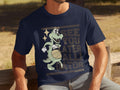 A man sits on a wooden bench in casual attire, wearing a navy blue Garment Graphics T-shirt featuring a cartoon alligator with a basketball and the playful motto, SEE YOU LATER ALLIGATOR, against a blurred outdoor scene.