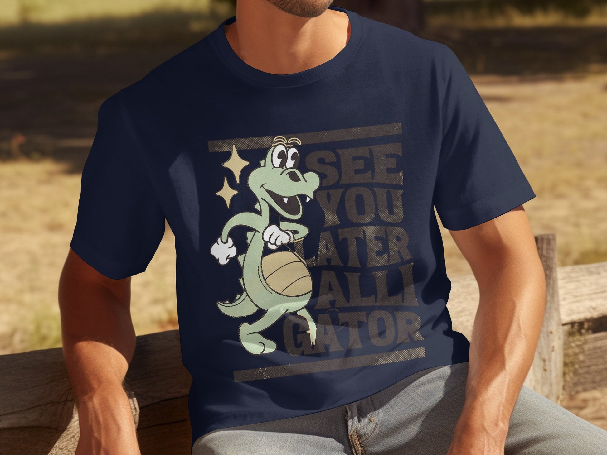 A man sits on a wooden bench in casual attire, wearing a navy blue Garment Graphics T-shirt featuring a cartoon alligator with a basketball and the playful motto, SEE YOU LATER ALLIGATOR, against a blurred outdoor scene.