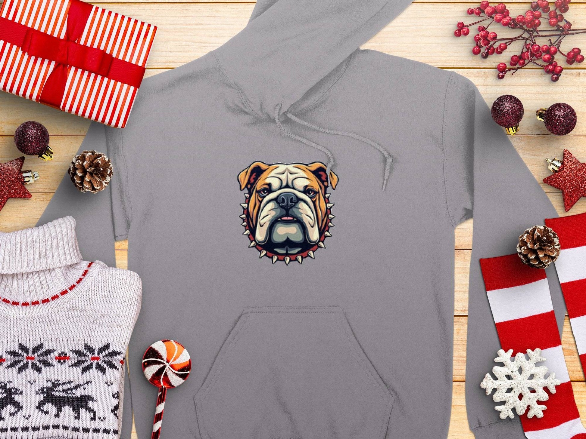 Garment Graphics presents a bold gray hoodie featuring a bulldogs face with a spiked collar, surrounded by festive wrapped gifts, pinecones, and Christmas ornaments—perfect for adding holiday cheer against its wooden background setting.