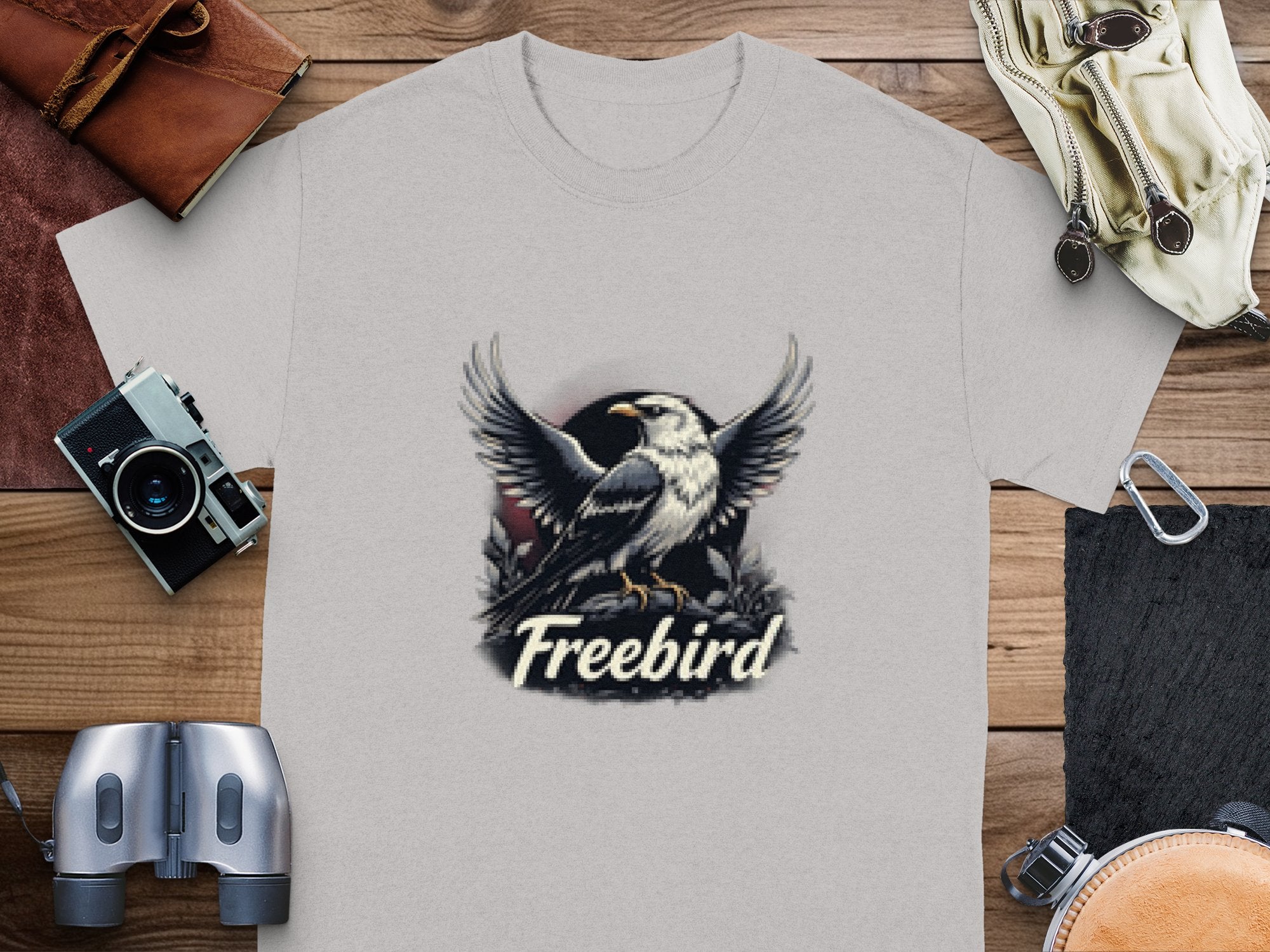 The Garment Graphics Freebird Eagle T-Shirt, made of 100% cotton, displays an eagle with outstretched wings and Freebird underneath. Its styled with a camera, binoculars, leather wallet, and canvas bag on a rustic wooden surface.