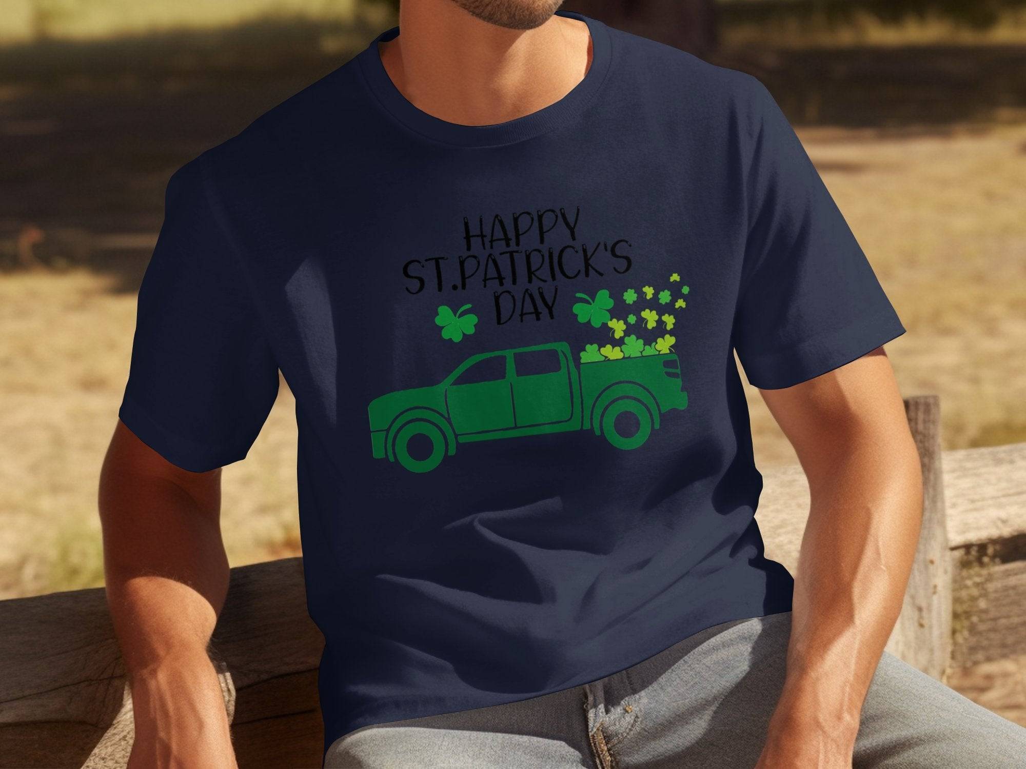 A person sits on a wooden bench wearing a navy blue Garment Graphics T-shirt featuring a green pickup truck adorned with shamrocks and the phrase Happy St. Patricks Day, celebrating Irish pride. In the background, grass and shadows subtly move in harmony.