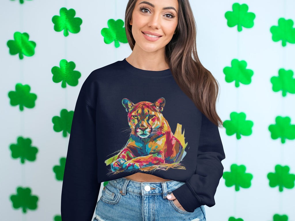 A woman in a Garment Graphics navy sweatshirt with a vibrant big cat design sits against green shamrock decorations. She has long brown hair and wears denim shorts, highlighting her unique style.