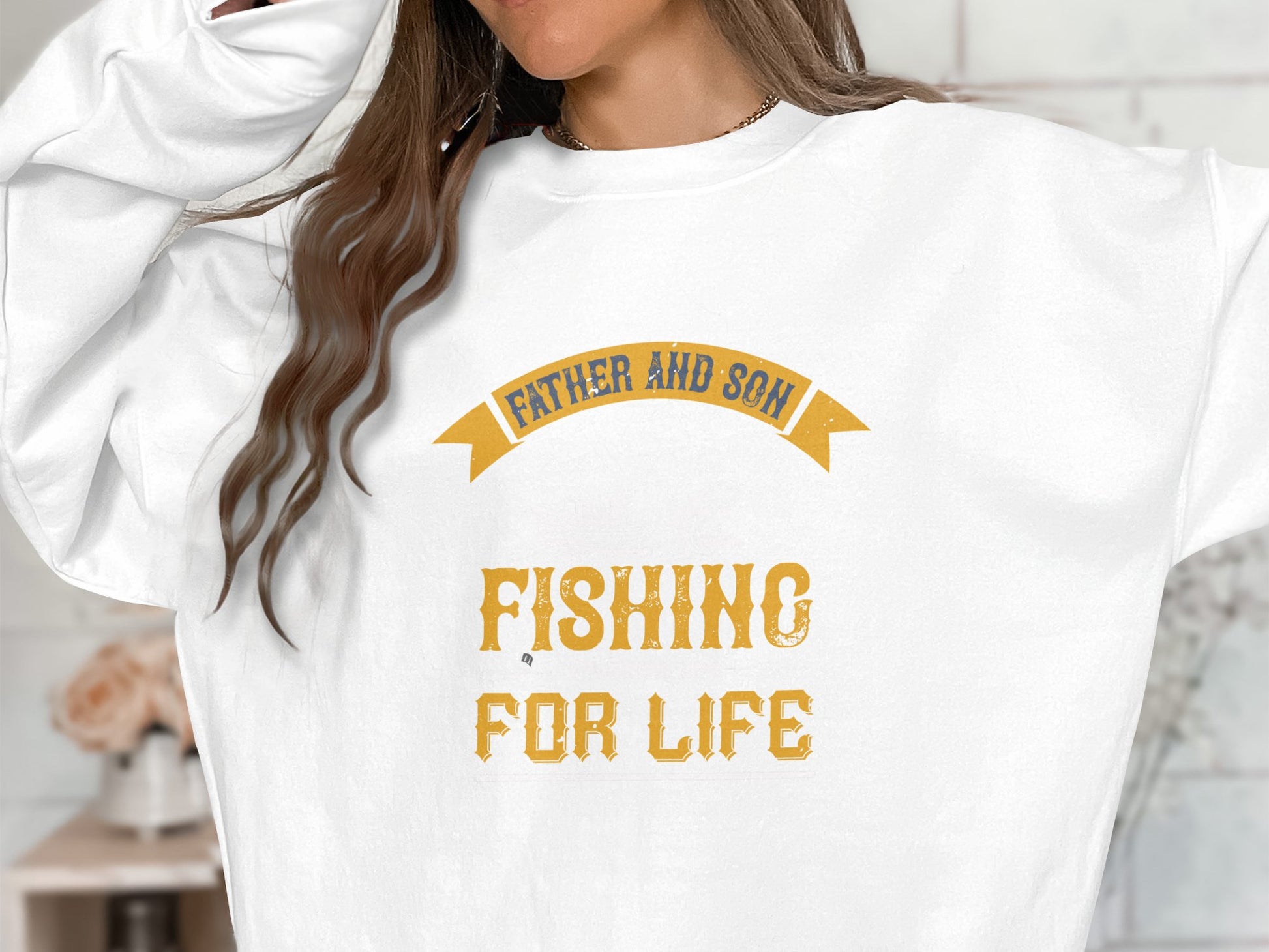 A person wearing a white Garment Graphics sweatshirt, featuring yellow text Father and Son Fishing for Life, stands with their long brown hair flowing. The softly blurred background conveys the serene essence of a father-son fishing trip.