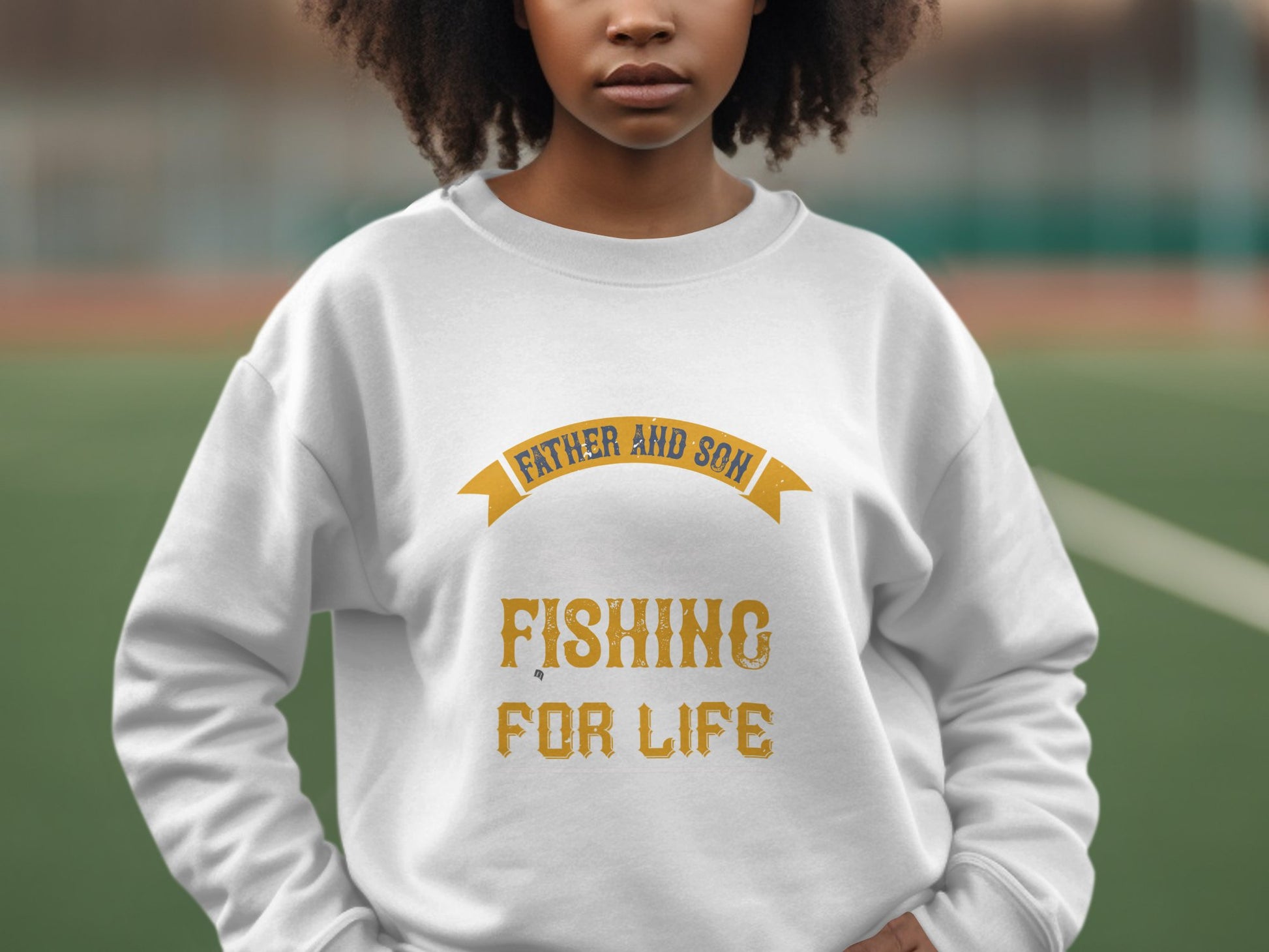 A person wears a white Garment Graphics sweatshirt featuring the phrase Father and Son Fishing for Life in bright yellow, set against a backdrop of blurred greenery and the edge of a sports field.