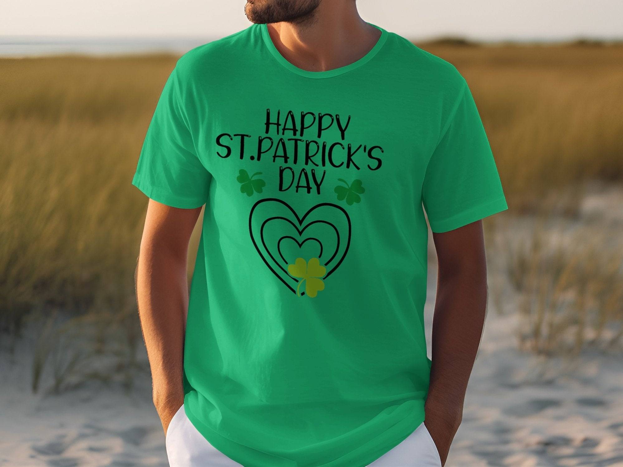 A person stands outdoors in a bright green Garment Graphics T-shirt with HAPPY ST. PATRICKS DAY in black, adorned with clovers and hearts in black and gold, adding a festive touch. Tall grasses and sandy terrain are seen behind them.