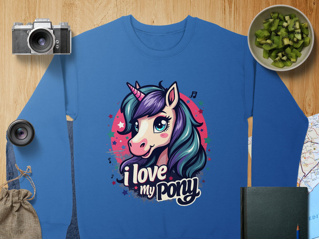 A blue graphic sweatshirt by Garment Graphics features a vibrant pony-unicorn design with the text I love my pony, accompanied by artful background elements: a camera, lens, sliced kiwi in a bowl, burlap bag, map, and dark green notebook on wood.