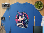 A blue graphic sweatshirt by Garment Graphics features a vibrant pony-unicorn design with the text I love my pony, accompanied by artful background elements: a camera, lens, sliced kiwi in a bowl, burlap bag, map, and dark green notebook on wood.