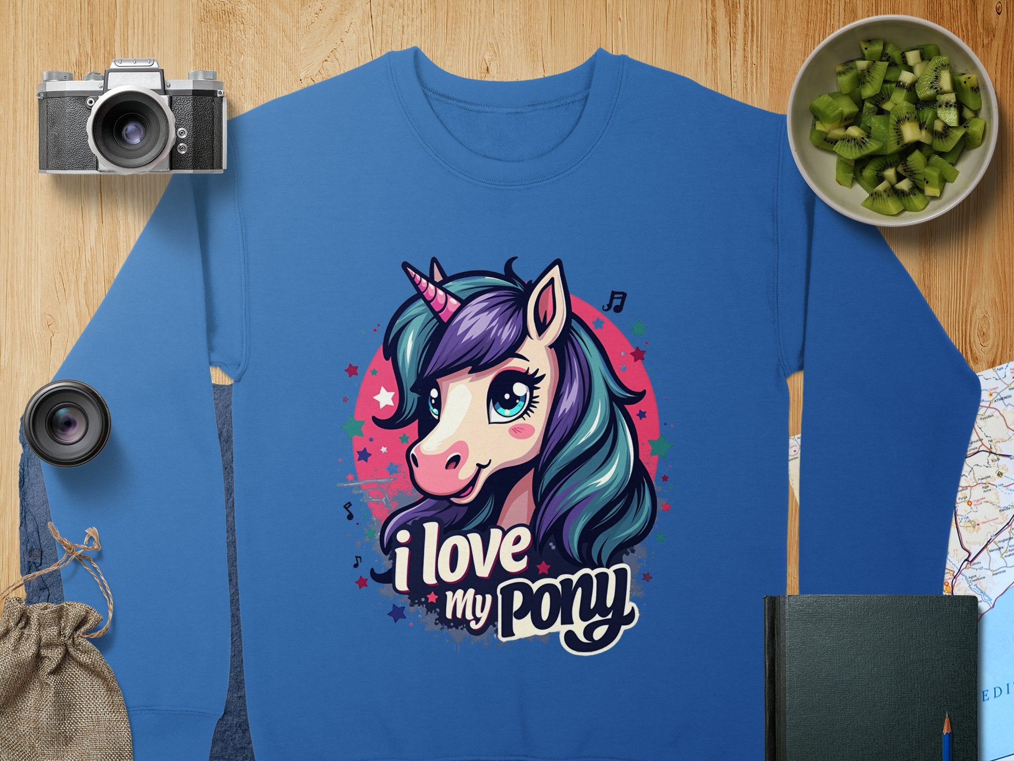 A blue graphic sweatshirt by Garment Graphics features a vibrant pony-unicorn design with the text I love my pony, accompanied by artful background elements: a camera, lens, sliced kiwi in a bowl, burlap bag, map, and dark green notebook on wood.