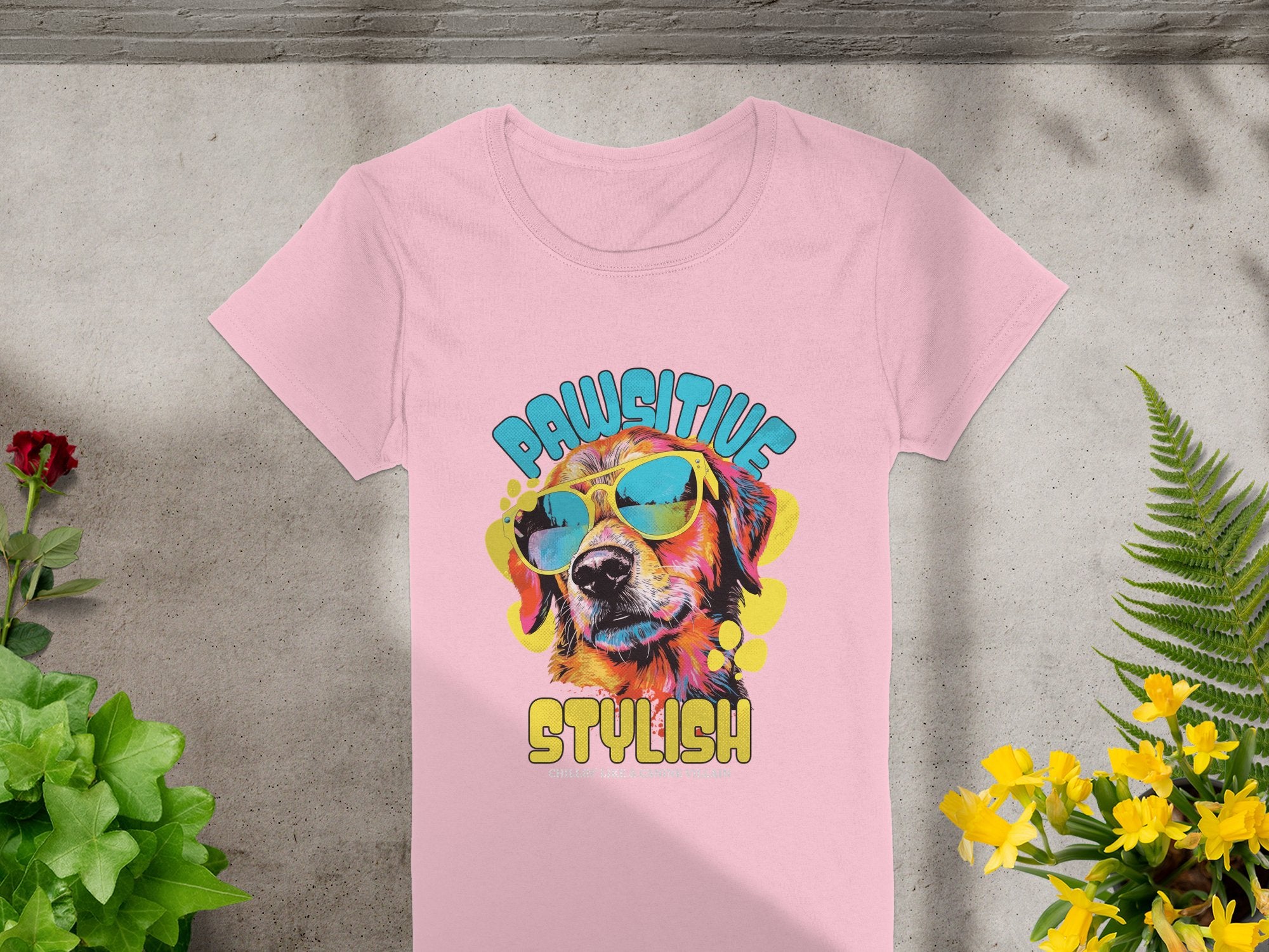 A pink Womens T-Shirt from Garment Graphics features a vibrant dog with sunglasses. PAWSITIVE is printed above and STYLISH below. Its photographed on concrete with small plants and flowers.
