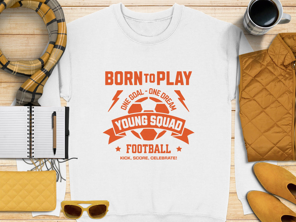 The Garment Graphics white t-shirt sports a vibrant orange soccer ball and Born to Play Young Squad Football print. Perfect with accessories like a scarf, mug, jacket, notebook, pen, sunglasses, and purse on wood surface. Unisex sizing ideal for young squads.