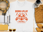 The Garment Graphics white t-shirt sports a vibrant orange soccer ball and Born to Play Young Squad Football print. Perfect with accessories like a scarf, mug, jacket, notebook, pen, sunglasses, and purse on wood surface. Unisex sizing ideal for young squads.