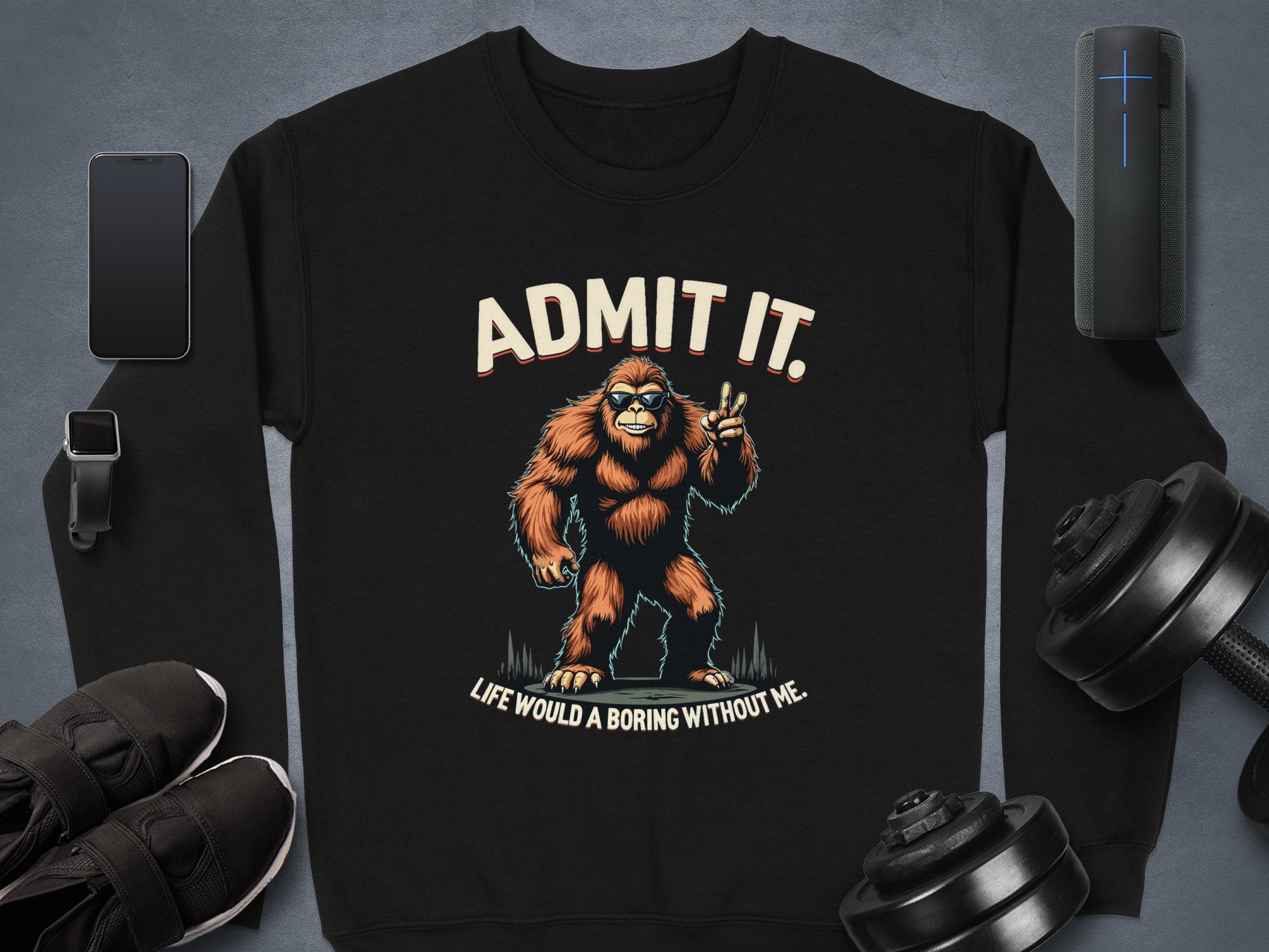 A black Garment Graphics sweatshirt with a striking Bigfoot graphic and the words, Admit It. Life Would Be Boring Without Me, lies beside a phone, smartwatch, sneakers, wireless speaker, and adjustable dumbbells on gray—an essential addition to any casual wardrobe.