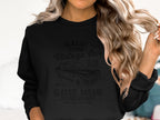 A person with shoulder-length wavy blonde hair wears a black sweatshirt from Garment Graphics, featuring a vintage car graphic and classic car club text. They hold a hair strand, embodying Classic London Citys timeless charm.