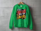 The Garment Graphics green sweatshirt on a wooden hanger features Its a Good Day to Laugh in vibrant fonts against a gray concrete wall. This Graphic Art Sweatshirt is available in unisex sizing for universal appeal and comfort.