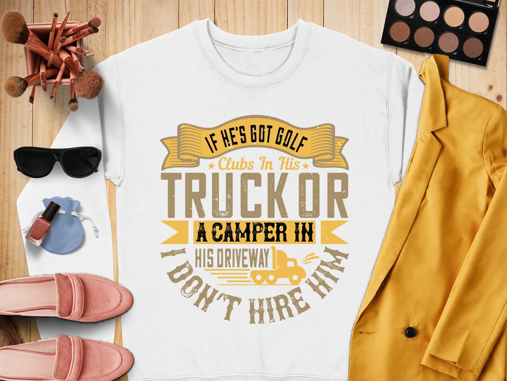 A flat lay displays a Garment Graphics sweatshirt with the text, If hes got golf clubs in his truck or a camper in his driveway, I dont hire him, alongside a mustard jacket, pink shoes, sunglasses, a watch, and makeup products on a wooden surface.