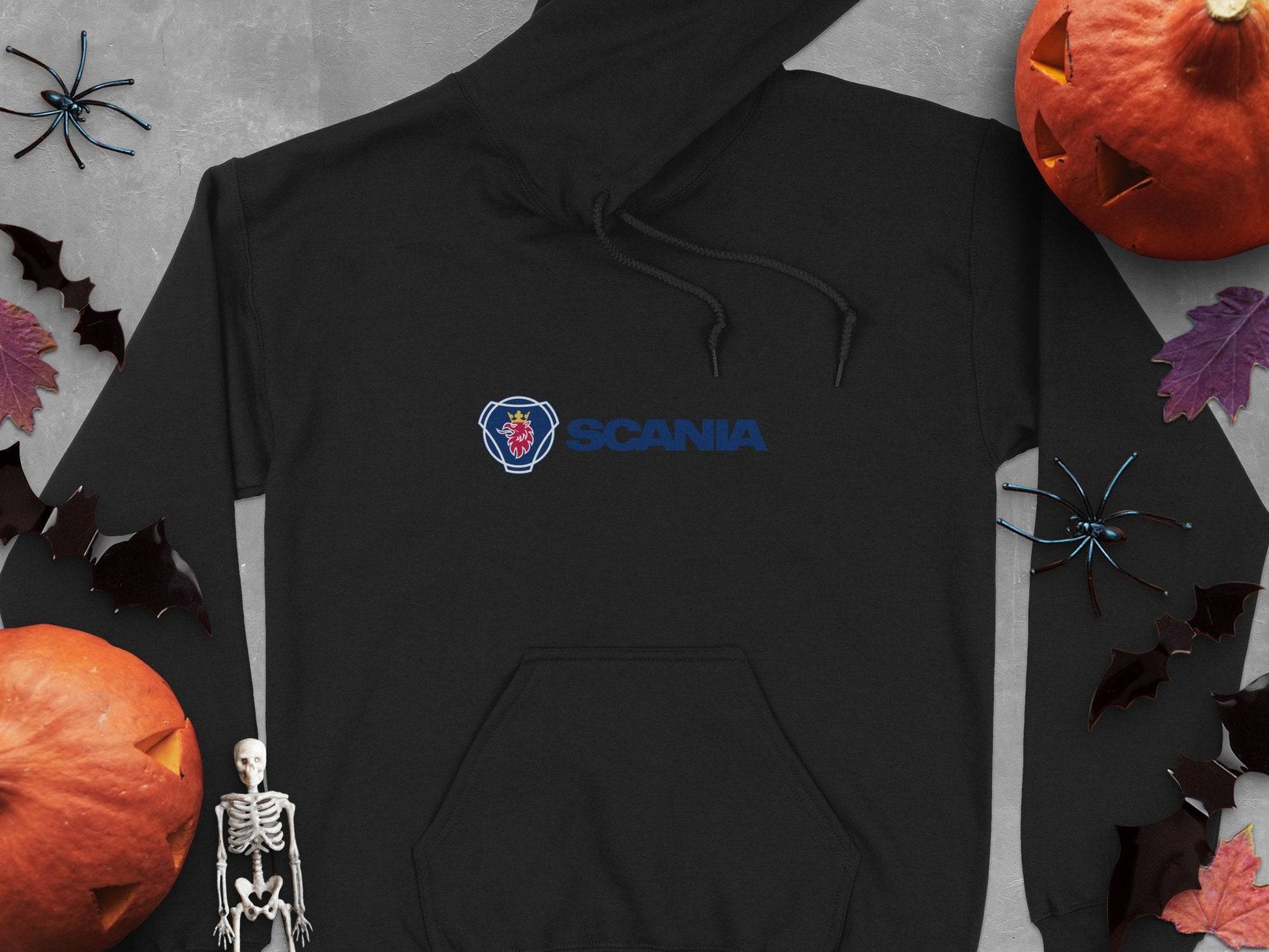 The Garment Graphics black hoodie proudly displays the blue Scania logo with a red emblem. Surrounded by Halloween decor like pumpkins, bats, spiders, and a skeleton on a gray backdrop, its ideal for fashion enthusiasts who appreciate unique style pieces.