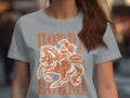 A person wears a Garment Graphics Rodeo T-shirt with a cowboy on horseback and Howdy Rodeo in bold orange text. Designed for womens sizing, it also features a Not a Beginner badge. The background is artfully blurred.