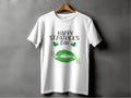The Garment Graphics T-shirt showcases a festive Happy St. Patricks Day message with green lips and clovers. Hanging against a plain gray wall, it perfectly blends style and celebration.