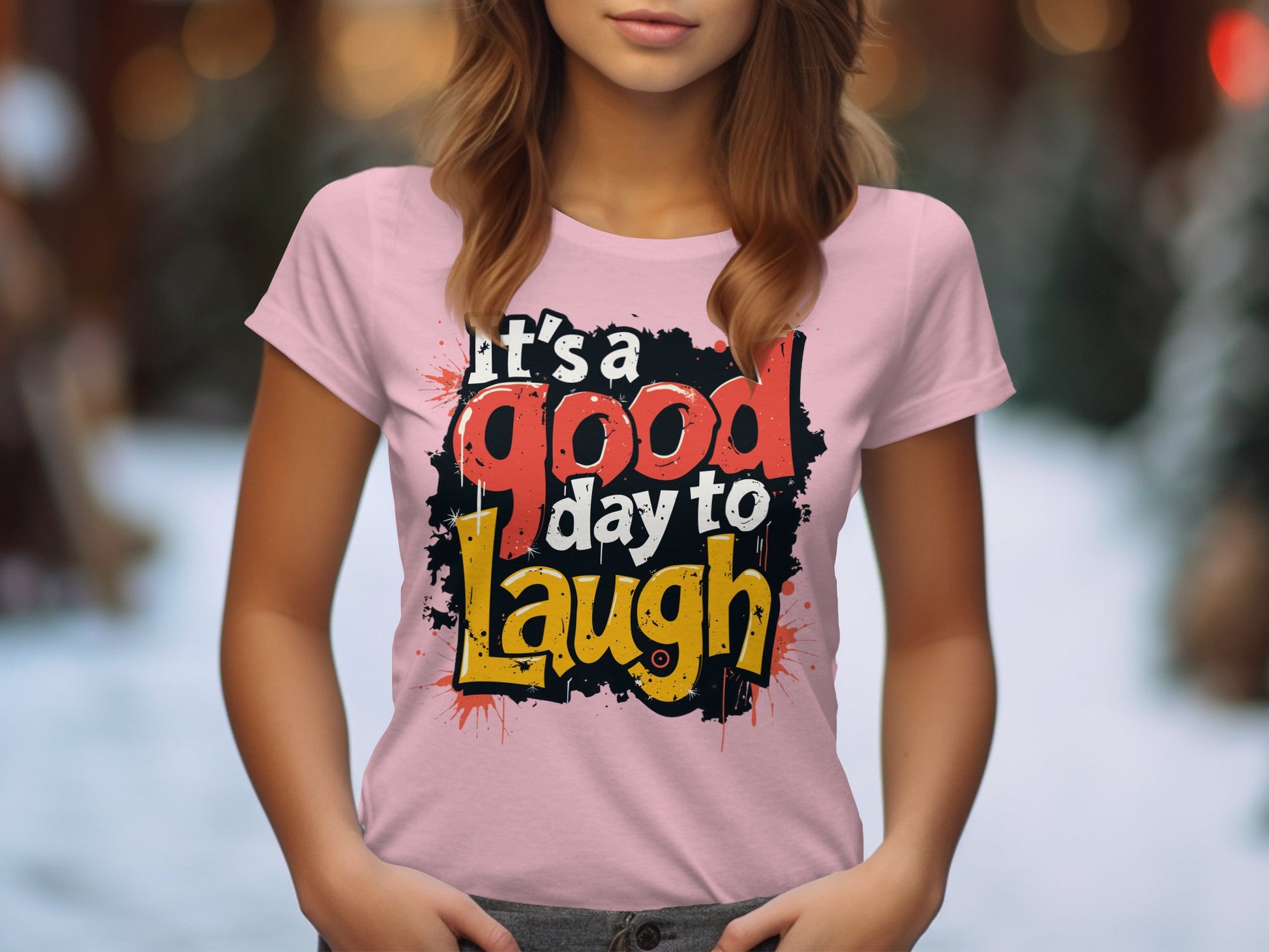 A person wears a semi-fitted pink Garment Graphics T-shirt with Its a good day to laugh in bold, colorful print. The blurred background of lights and greenery enhances the positive vibe.