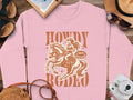 Garment Graphics presents a pink sweatshirt with a vivid cowgirl-on-horse graphic and Howdy Rodeo slogan. Featuring items like a camera, hat, and sunglasses alongside the phrase Not a beginner, its perfect for rodeo enthusiasts!.