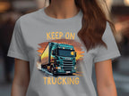 A person with long brown hair wears a light gray shirt from Garment Graphics featuring a bold truck graphic driving at sunset and the phrase KEEP ON TRUCKING, making it perfect for trucking enthusiasts.