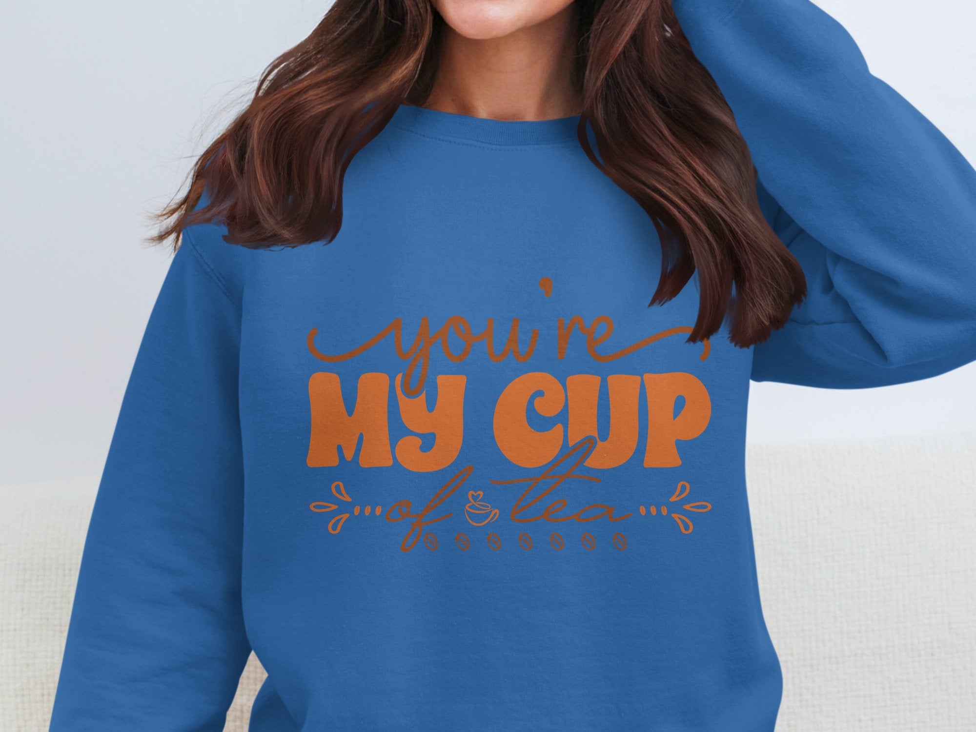 Someone is wearing a cozy unisex-sized blue sweatshirt from Garment Graphics, featuring Youre My Cup of Tea in vibrant orange. Their face is not visible, and they have long brown hair.