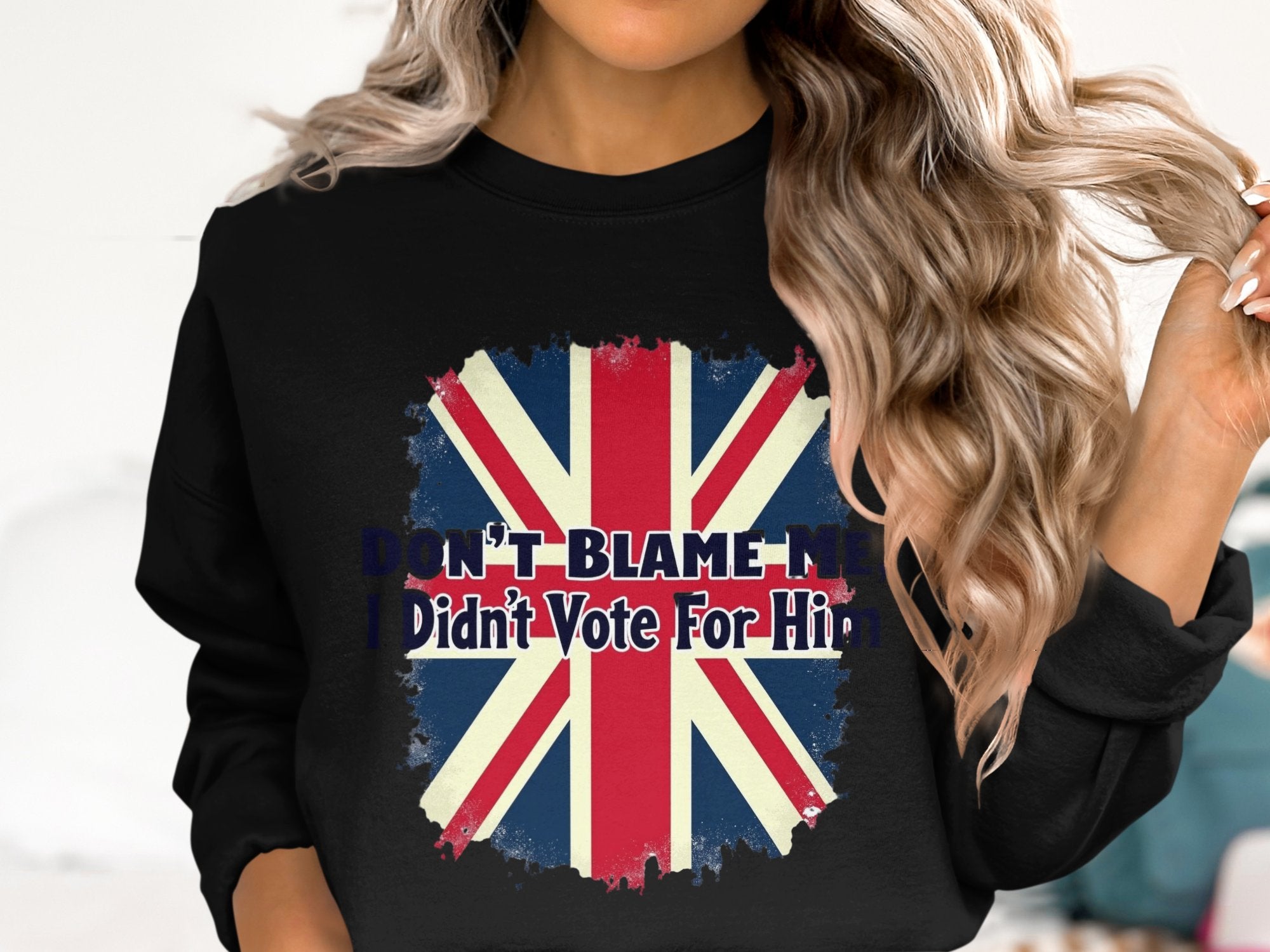 A person with long, wavy hair sports a black Garment Graphics sweatshirt displaying a faded Union Jack and the slogan Dont Blame Me, I Didnt Vote For Him. Unisex sizing offers comfort for everyone.