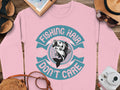 Garment Graphics offers a pink fishing sweatshirt with a fish graphic and the text Fishing Hair Dont Care in teal and white, styled alongside accessories like a straw hat, sunglasses, notepad, camera, and shoes on a wooden surface for the fishing enthusiast.