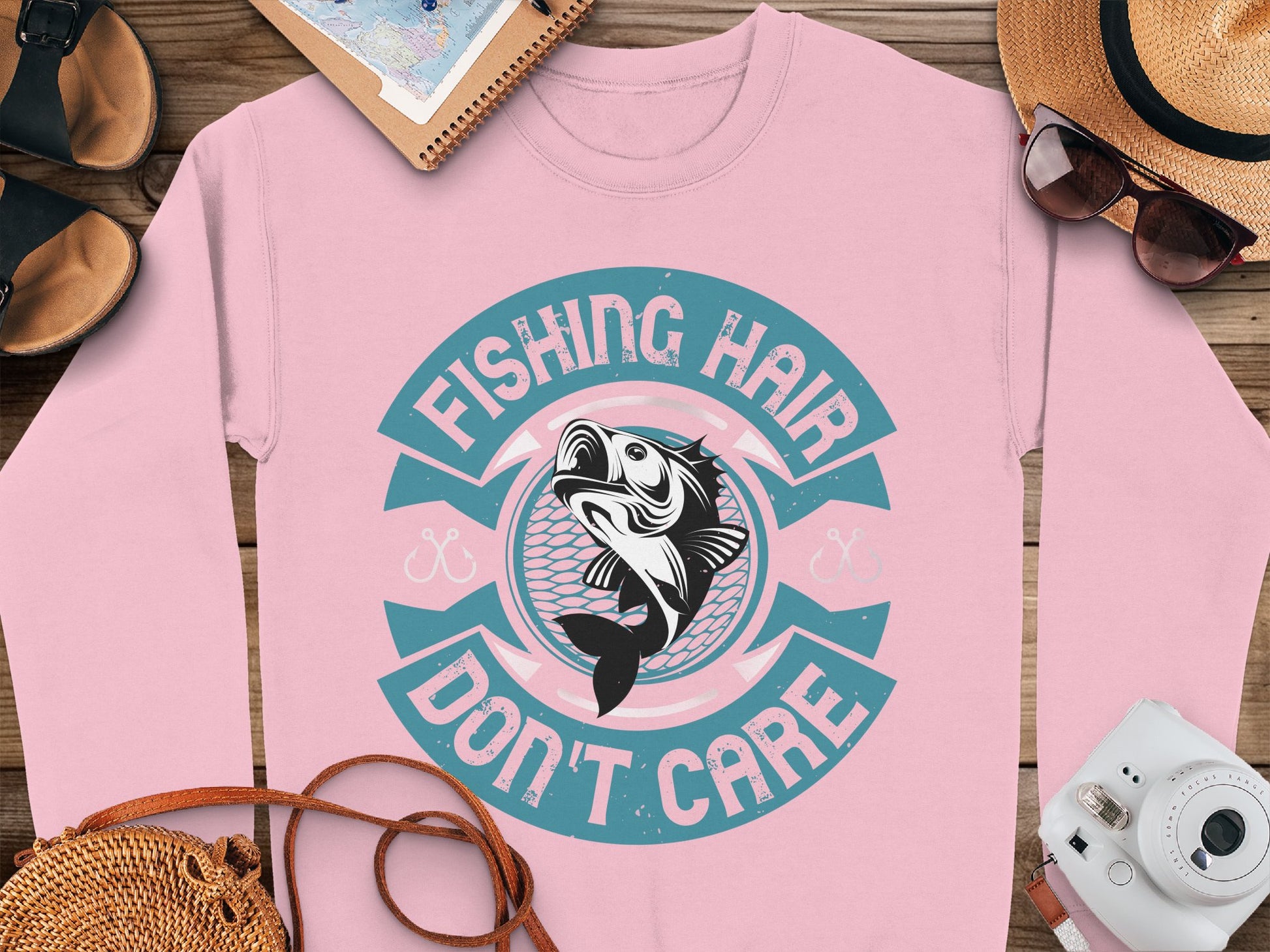 Garment Graphics offers a pink fishing sweatshirt with a fish graphic and the text Fishing Hair Dont Care in teal and white, styled alongside accessories like a straw hat, sunglasses, notepad, camera, and shoes on a wooden surface for the fishing enthusiast.