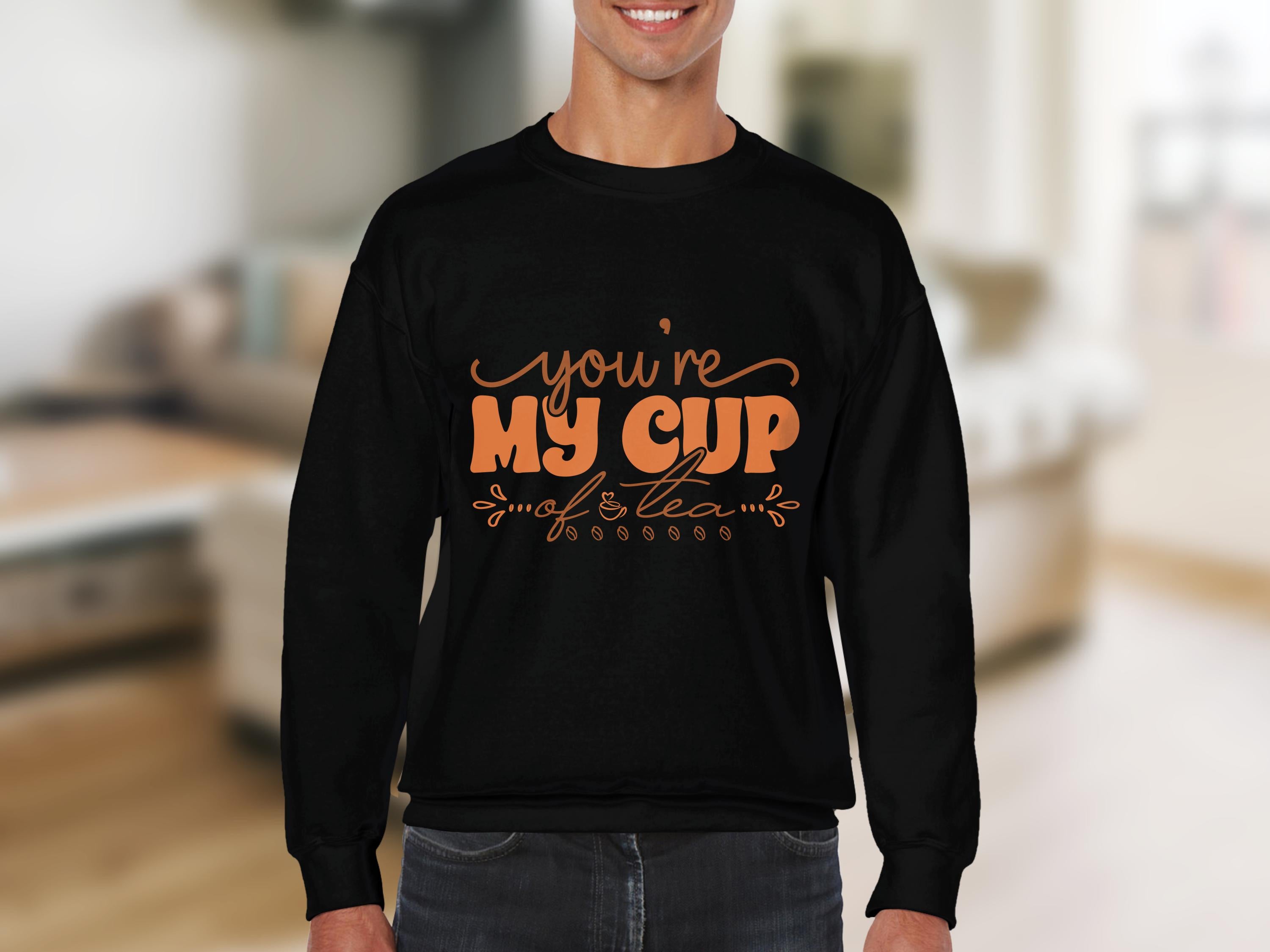 A tea enthusiast wearing a cozy black Garment Graphics sweatshirt with bold orange youre my cup of tea lettering stands indoors. The softly blurred background reveals a room with neutral-colored furniture.